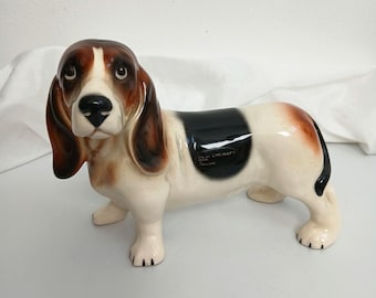 Puede incluir: Figura de perro basset hound de cerámica con acabado brillante. El perro tiene marcas marrones y blancas en la cabeza y las orejas, una mancha negra en la espalda y manchas marrones en la parte trasera. La figura está de pie sobre sus cuatro patas.