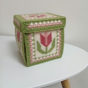 Puede incluir: Caja cuadrada hecha a mano con tapa, con un diseño de punto de cruz. La caja es principalmente verde, con detalles rosas y blancos. Cada lado muestra un motivo de tulipán rojo. La caja está sobre una mesa blanca.