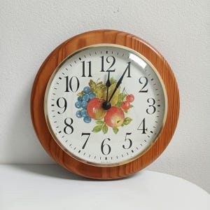 May include: A round wooden wall clock with a light-coloured face. The clock face features black numerals, black hands, and a decorative fruit design with apples, grapes, and cherries. The clock has a gold-coloured bezel.