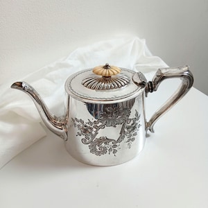 Puede incluir: Tetera plateada con boquilla y asa curvadas. La tapa tiene un pomo decorativo de color marfil. Grabados florales adornan el cuerpo de la tetera. La tetera es probablemente de plata o material plateado.