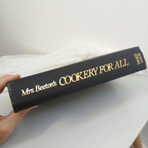Peut inclure: Un livre relié noir avec des lettres dorées sur le dos indiquant Mrs Beeton's COOKERY FOR ALL et WARD LOCK. Le livre est positionné horizontalement, mettant clairement en évidence le titre et les détails de l'éditeur sur la couverture en tissu.