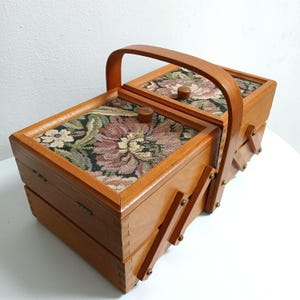 Può includere: Una scatola da cucito in legno con coperchio incernierato e manico. La scatola è marrone chiaro con un disegno arazzo floreale sul coperchio. La scatola ha un cassetto e un meccanismo pieghevole sul lato.