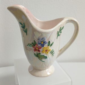 May include: Cream ceramic creamer with a floral design. The creamer has a pink interior and a handle. The floral design includes blue, yellow, and pink flowers with green leaves. The creamer sits on a small pedestal base.