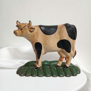 以下が含まれることがあります： ベージュの体、黒い斑点、緑色のベースが特徴的な装飾用の牛の置物。牛には角があり、緑色のテクスチャベースの上に立っています。この置物は頑丈な素材でできており、室内装飾用である可能性があります。