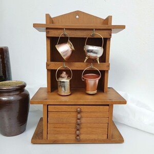 May include: A wooden wall-mounted rack featuring a top shelf with four metal hooks holding miniature copper and silver-toned vessels, including a pot, a bucket, and a canister. Below the shelf is a stack of six wooden drawers with round knobs.