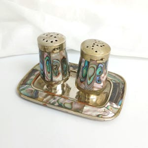 May include: A vintage salt and pepper shaker set. The shakers and tray are made of brass and inlaid with iridescent abalone shell. The shakers have a classic cylindrical shape with perforated tops. The rectangular tray has a matching abalone shell inlay.