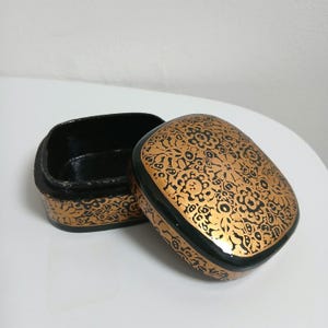 May include: An open, square-shaped box with a lid. The lid and sides are black with a gold floral pattern. The interior of the box is black. The box is sitting on a white surface.