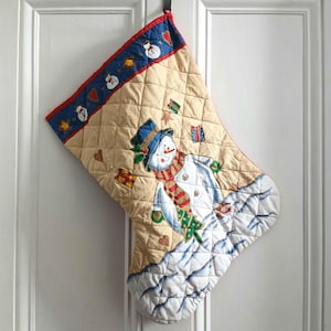 May include: A quilted Christmas stocking in beige with a blue cuff featuring snowmen and hearts. The main body displays a snowman wearing a blue top hat and a red and green striped scarf, surrounded by small gift boxes and script text.