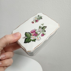 以下が含まれることがあります： イチゴのデザインが施された蓋付きの白いセラミック製の小物入れ。 イチゴはピンクと赤で、緑の葉と白い花が付いています。 ボックスには金のトリムが付いています。