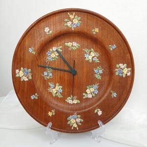 May include: A round wooden wall clock with a rich brown grain finish. The clock face is decorated with small, raised floral clusters in shades of yellow, blue, and pink. It features two thin, black metal hands centred on the circular wooden dial.