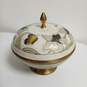 Może przedstawiać: Ozdobna, ceramiczna miska z pokrywką, złotą podstawą i obramowaniem. Kremowa pokrywka ma motyw motyla i kwiatowy w odcieniach brązu, czerni i złota. Zakończenie jest w kolorze złotym.