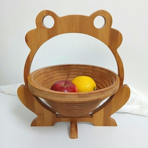 Peut inclure: Un panier à fruits en bambou en forme de grenouille, avec un bol rond et une poignée. Il contient une pomme rouge et un citron jaune. Le panier est en bambou brun clair.
