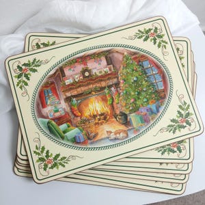 May include: A set of rectangular placemats featuring a cream-colored border with green holly leaf illustrations. The central oval illustration depicts a cozy living room with a lit fireplace, a decorated Christmas tree, and a sleeping cat.