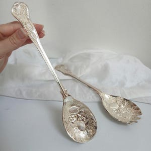 May include: A pair of silver-toned metal salad serving utensils featuring long, slender handles with ornate shell-like detailing at the top. The bowls of the spoons are embossed with detailed fruit motifs, and one spoon features a three-pronged fork edge.