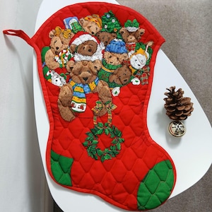 May include: A quilted red Christmas stocking featuring a printed design of multiple brown teddy bears wearing winter scarves and hats, a green Christmas tree, and a green holly wreath. The stocking has green quilted accents at the toe and heel.