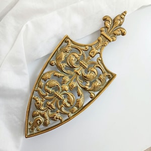 Peut inclure: Un dessous de plat doré orné d'un motif de fleur de lys. Le dessous de plat présente un motif ajouré de feuilles et de motifs floraux tourbillonnants. La pièce décorative est en forme de bouclier.