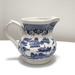 Puede incluir: Jarra de cerámica blanca con un diseño azul de patrón Willow. La jarra tiene un asa curva y un pico vertedor. El diseño azul y blanco representa un paisaje con edificios, árboles y un barco.