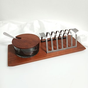 May include: A rectangular wooden serving tray featuring a circular glass container with a matching wooden lid and a small metal spoon, alongside a metal toast rack with six vertical slots for holding slices of bread in an upright position.