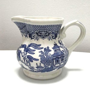Può includere: Una lattiera in ceramica bianca con un motivo blu a trasferimento raffigurante una pagoda, alberi di salice e bordi geometrici. La lattiera presenta un manico curvo, un ampio beccuccio e un corpo arrotondato con bordo e base svasati.