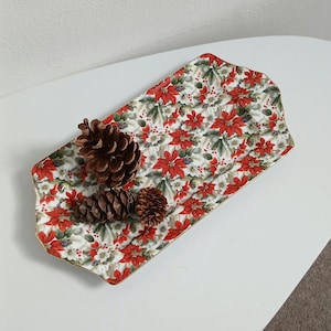 May include: A rectangular serving tray with a floral poinsettia pattern in red, white, and green. Three brown pine cones rest on the tray. The tray has a gold trim and is placed on a white surface.