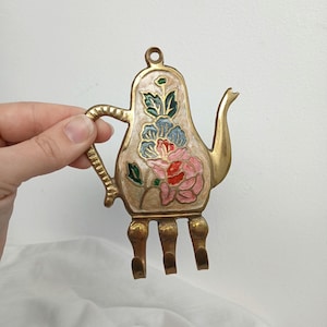 May include: A brass teapot-shaped wall hook with three hooks. The teapot is painted with a floral design in pink, blue, and green. The handle and spout are detailed. The hook is held by a hand.