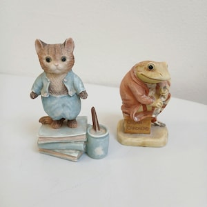 Puede incluir: Dos figuras de cerámica: un gato con un atuendo azul de pie sobre libros y una rana con un abrigo que sostiene un letrero que dice "Cream Crackers". El gato tiene pelaje marrón y canela, y la rana lleva un abrigo rojo.