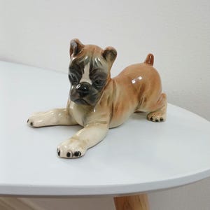 Op de afbeelding: Een keramieken Boxer hond beeldje in een ontspannen pose. De hond heeft een bruin lichaam met een wit en zwart gezicht, en zwarte poten. Het beeldje staat op een wit oppervlak.