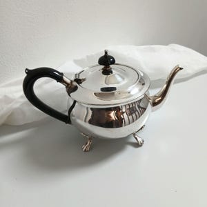 Puede incluir: Una tetera plateada con asa negra y pomo negro en la tapa. La tetera tiene un pico curvo y se apoya en tres pequeños pies. La superficie brillante refleja el entorno.