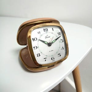 May include: A vintage Starlet alarm clock with a gold-tone frame and a light pink case. The clock face is white with black numerals and hands, and the word "ALARM" is printed on the face. The clock is open, revealing the face.