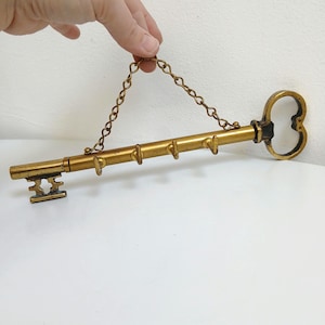 以下が含まれることがあります： 金色の金属で作られた、ヴィンテージスタイルの鍵型の壁掛けフック。 鍵は装飾的な頭部、シャフトに沿った一連の小さなフック、および吊り下げ用のチェーンを備えています。 白い背景に対して、手で鍵が持ち上げられています。