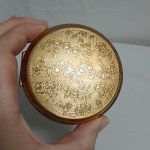 May include: A round, gold-toned compact with a floral design. The lid features an embossed pattern of flowers and leaves. The compact is held in a hand, showcasing its size and detail. The compact has a hinged opening and a brushed metal finish.