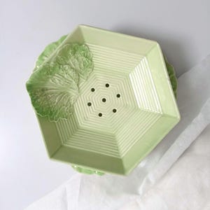 Può includere: Un piatto da portata in ceramica verde chiaro con una forma esagonale unica. Il piatto presenta un design strutturato con un motivo centrale di linee radiali e sei piccoli fori. Accenti decorativi di foglie di cavolo sul lato.