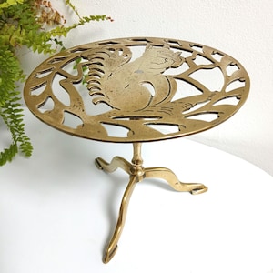 May include: A vintage brass trivet featuring a squirrel motif. The round trivet has a cut-out squirrel and branch design, supported by three curved legs. The brass has a warm, aged patina, typical of vintage items.