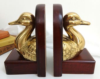 May include: A pair of bookends featuring polished brass duck figures mounted on dark brown wooden bases. Each duck has a smooth head and neck with a textured body. The bookends are positioned side by side, showcasing the metallic sheen and wood grain.