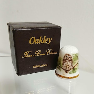 Puede incluir: Un dedal de porcelana blanca con un diseño pintado de un búho y una rama de pino, junto a una caja marrón con letras doradas que dicen "Oakley Fine Bone China ENGLAND".