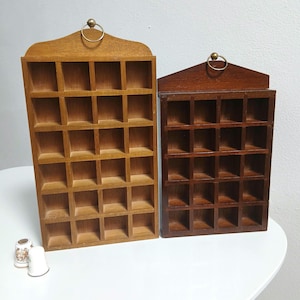 Puede incluir: Dos estanterías de madera con múltiples compartimentos cuadrados pequeños. Una estantería es de color marrón claro, la otra es de color marrón oscuro. Cada una tiene una parte superior decorativa con un anillo de latón para colgar. Las estanterías están vacías, listas para exhibir objetos pequeños.