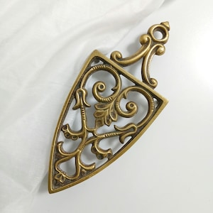 May include: A triangular brass trivet featuring an openwork design with scrolling leaf and vine motifs. The metal object has a pointed base and a handle at the top with a circular cutout for hanging, showcasing a metallic gold finish.