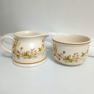 May include: A cream-colored ceramic creamer and sugar bowl set featuring a botanical pattern of green leaves, small white flowers, brown berries, and golden wheat stalks. Both pieces have a brown band near the base and a smooth, rounded shape.
