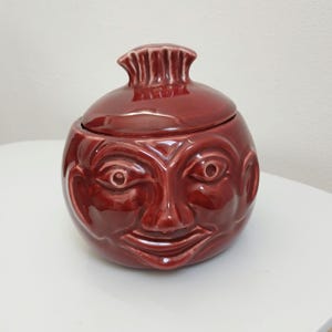 May include: A maroon ceramic jar with a lid, featuring a sculpted face design. The jar has a round body and a textured lid. The face has detailed eyes, a nose, and a smiling mouth. The jar is on a white surface.