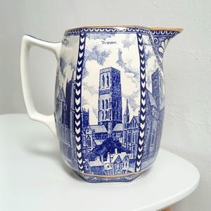 May include: A blue and white ceramic jug featuring a detailed illustration of Durham Cathedral, with the word "Durham" above the image. The jug has a gold rim and handle. The design is in a classic blue and white style.