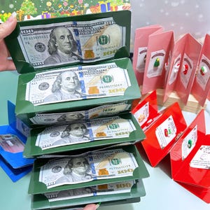 May include: A display of green gift boxes containing imitation $100 bills, alongside pink and red Christmas cards. The bills feature a portrait of Benjamin Franklin.