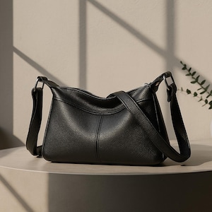 May include: A black leather shoulder bag with a textured finish. The bag has a rectangular shape with a curved top and a long adjustable strap. The bag is sitting on a light-colored surface.