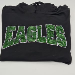Könnte beinhalten: Schwarzer Kapuzenpullover mit dem Wort "EAGLES" in großen, grünen Buchstaben im Strass-Stil auf der Vorderseite. Der Hoodie hat einen Kordelzug und ist ordentlich gefaltet.
