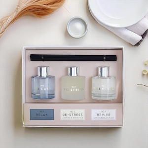 Op de afbeelding: Een doos met drie glazen aromatherapie diffusers met zilveren doppen. De flessen zijn gelabeld met "RELAX", "DE-STRESS" en "REVIVE". Elke fles bevat een vloeistof van een andere kleur. De set bevat zwarte diffuser sticks en labels met geurbeschrijvingen.