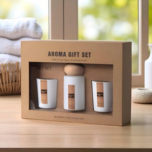 Op de afbeelding: Aroma cadeau set in een kartonnen doos. De set bevat een witte diffuser met een houten bovenkant, twee witte kaarsen en de tekst "AMBER ALLURE". De doos is gelabeld "AROMA GIFT SET".