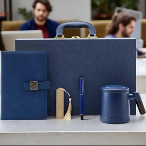 以下が含まれることがあります： ネイビーブルーのオフィス用品のコレクション。ブリーフケース、装飾的な留め金が付いたジャーナル、ペン、木製のハンドルが付いたマグカップ、タッセル付きのブックマークが含まれています。ブリーフケースには、金色のアクセントが付いたハンドルが付いています。