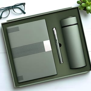 May include: A gift set in a green box, containing a notebook, pen, and insulated tumbler. The notebook has horizontal stripes and a silver clasp. The pen is silver, and the tumbler is cylindrical. The set is presented on a white wooden surface.