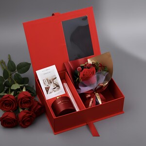 May include: A red gift box with a bouquet of red roses, a reed diffuser, and a card. The box has a window and a ribbon closure. The bouquet is wrapped in purple and gold paper with a ribbon that says "Eternal".