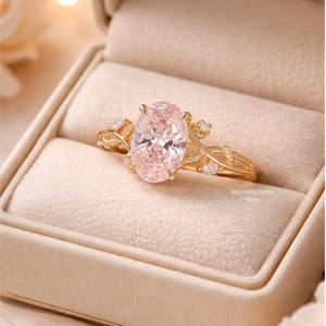 May include: A gold ring with a large, oval-cut pink gemstone, set in a delicate, vine-like design. The ring is displayed in a beige ring box, showcasing the intricate details of the jewelry. The ring is from the Gopinathji Gems Store.