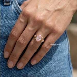 May include: A gold ring featuring two oval-cut pink gemstones. The ring has a delicate, ornate design with smaller accent stones. The ring is displayed on a hand, with a denim background.